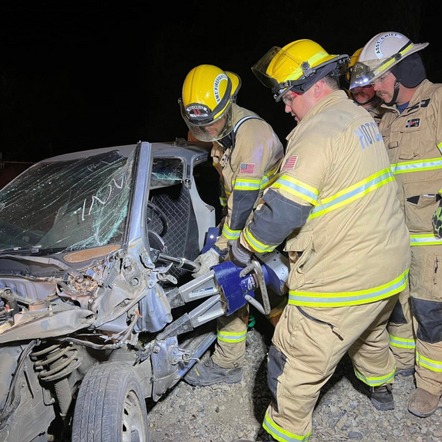 Firefighters in yellow helmets rescue a heavily damaged car driver at night.