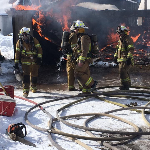 Firefighters in reflective gear battle a house fire amid smoke, flames, and hoses on snowy ground.