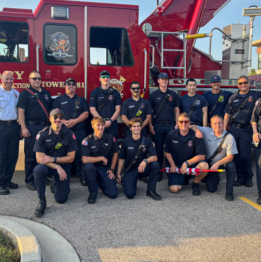 Group of firefighters posing in front of a red fire truck, wearing dark uniforms and sunglasses.