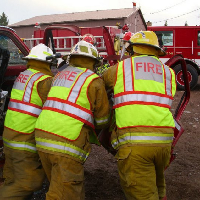 Three firefighters in yellow jackets and helmets crouch beside a red fire truck at a muddy scene.