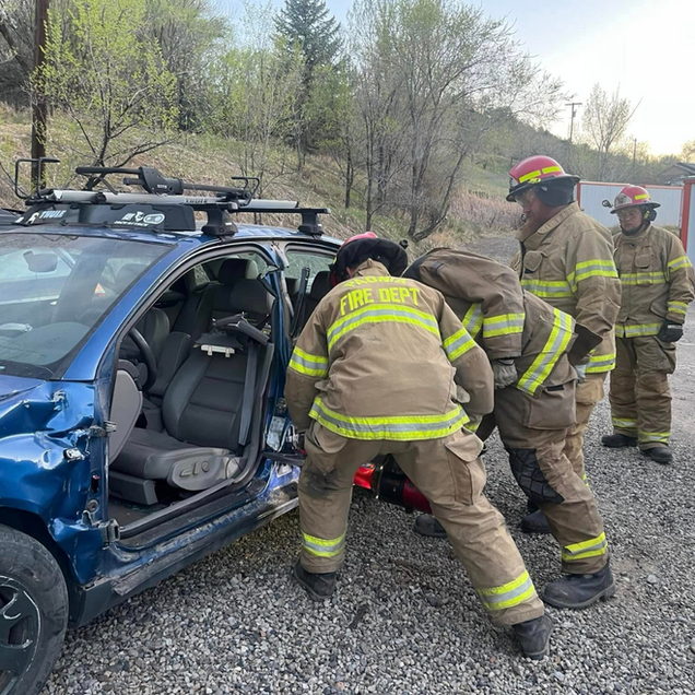 Firefighters in tan gear rescue someone from a blue SUV after a roadside crash.