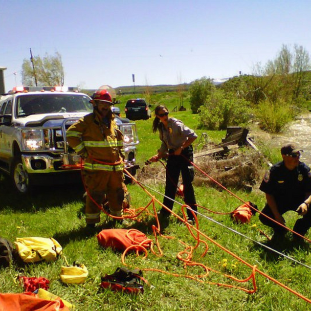 Firefighters and rescue gear beside a vehicle in a grassy roadside area, with tangled orange ropes on the ground