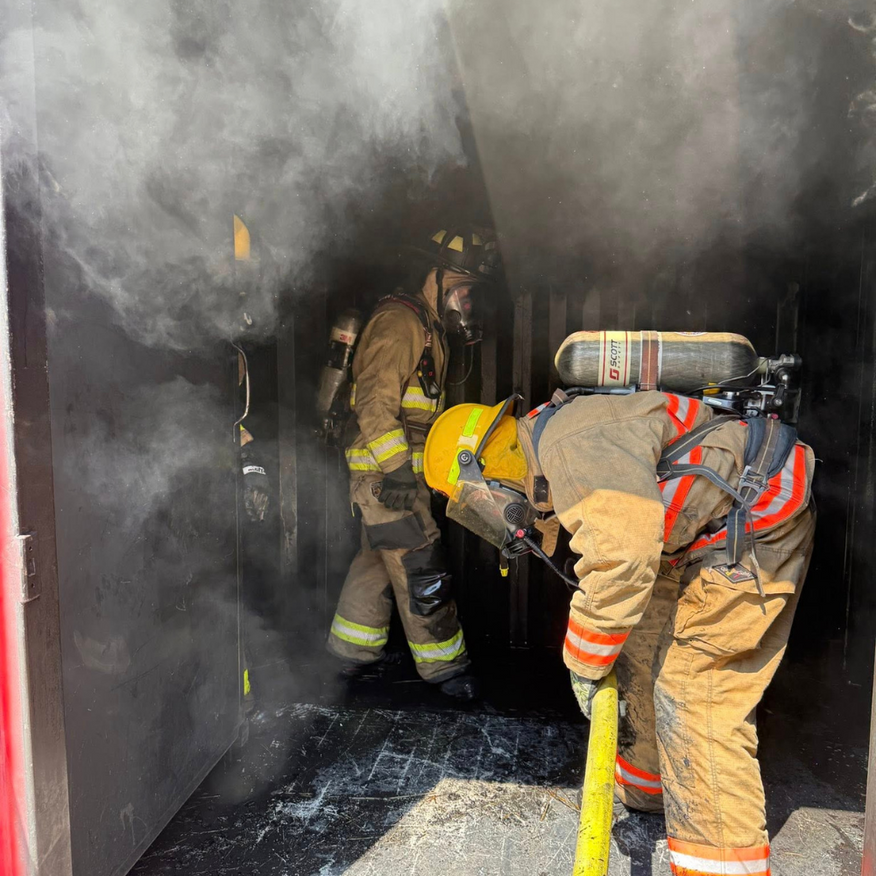 Firefighters in bunker gear inside a smoky structure, one spraying water.