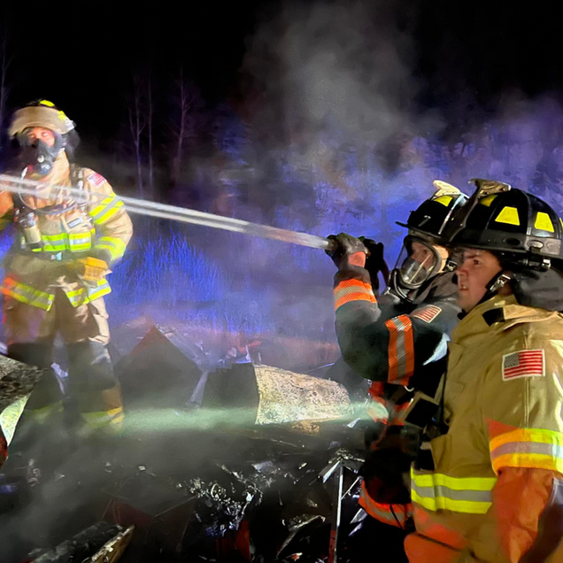 Firefighters in yellow gear spray water on a smoky nighttime fire scene
