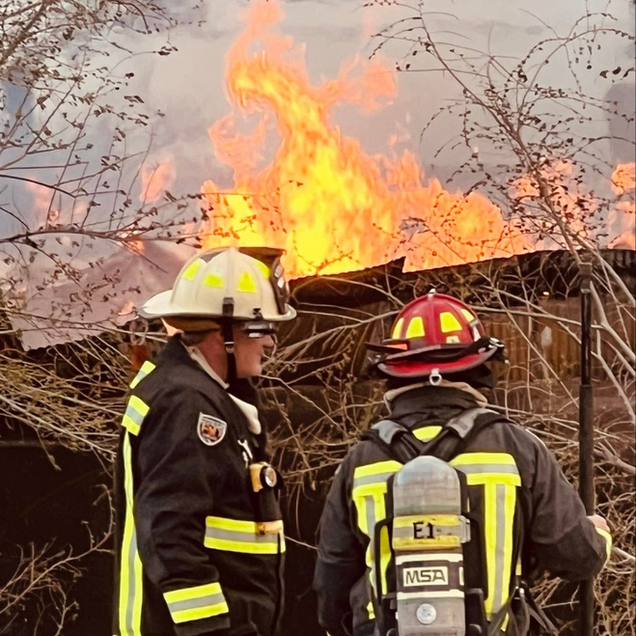 Two firefighters in reflective gear stand before a burning structure with flames and smoke.