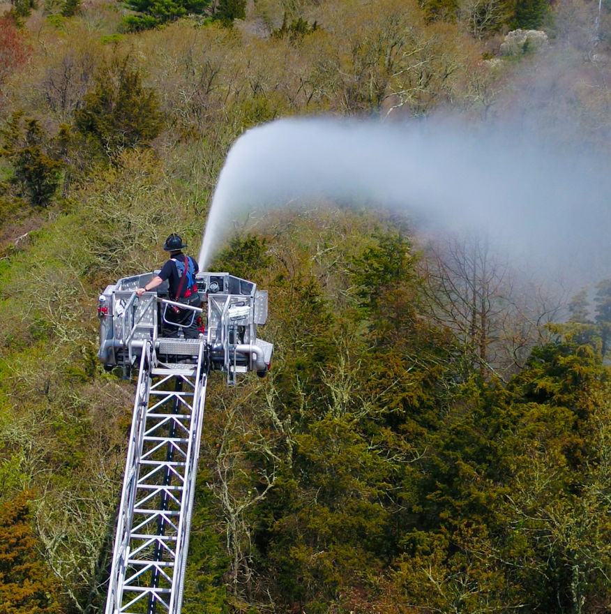 Firefighter on an extended ladder spraying water over trees at a wildfire scene