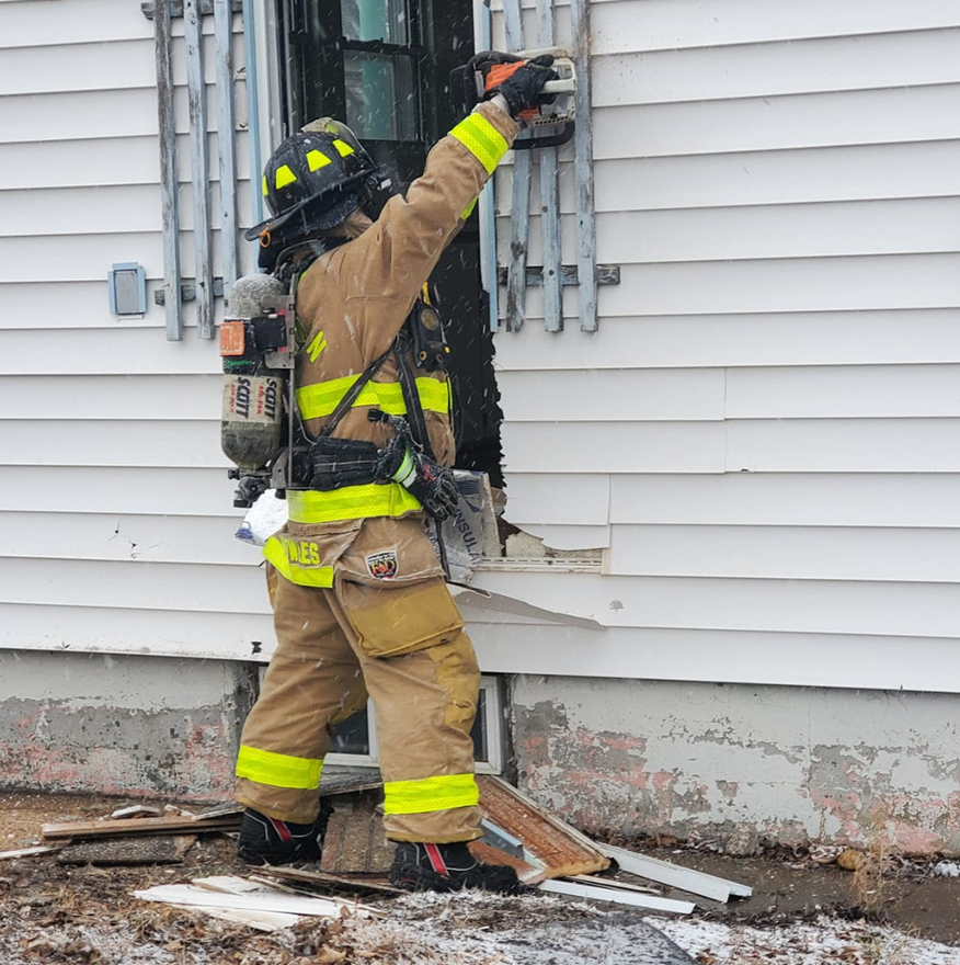 Firefighter in protective gear using a tool to breach a window in a white house.