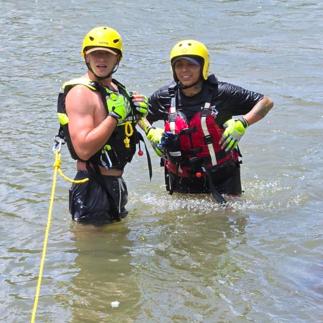 Two helmeted rescuers waist-deep in water, one holding a rope, both wearing life vests and gloves.