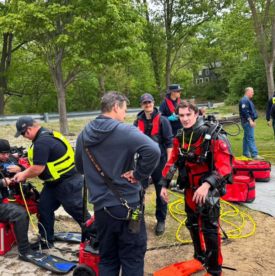 Diver team in wetsuits and red gear preparing equipment by a lakeshore on a sandy wooded shore
