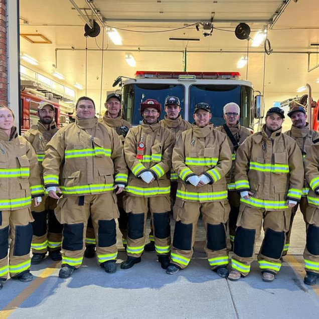 Firefighters in tan turnout gear standing in a fire station bay in front of a fire truck.