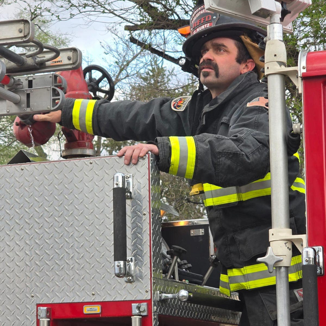 Firefighter in black gear directing from a fire truck ladder platform, with red equipment nearby.