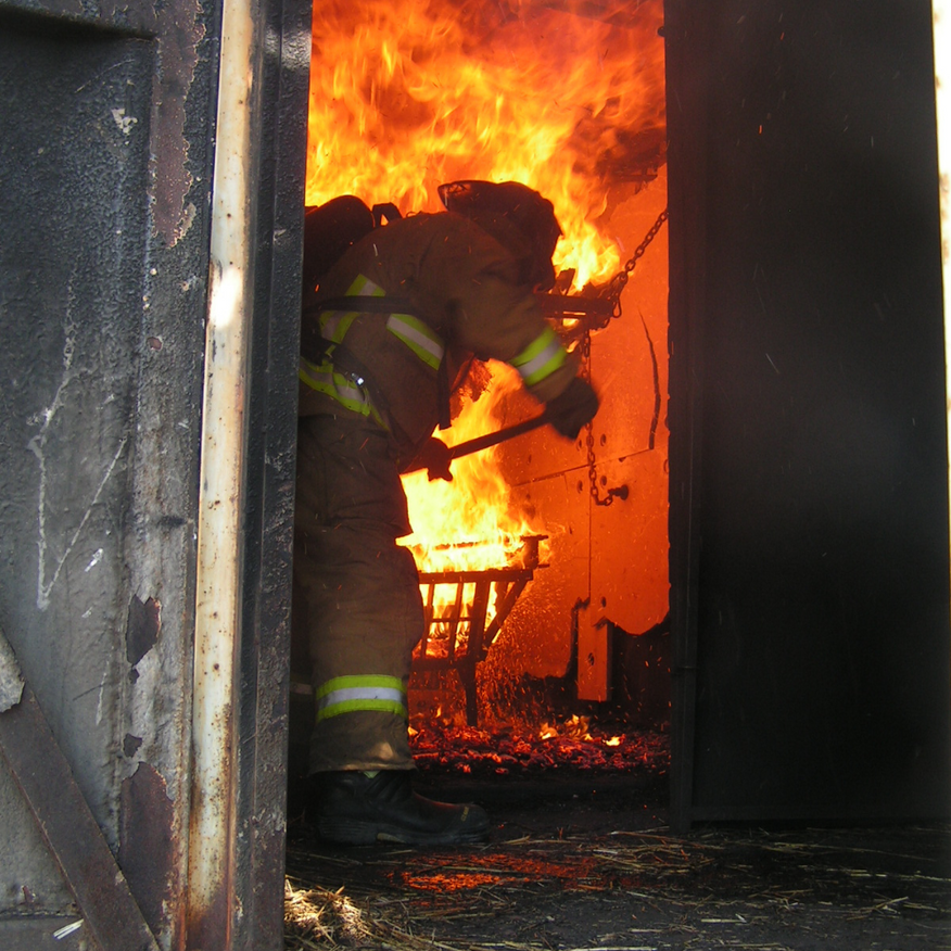 Firefighter in full gear battling a blaze inside a doorway, flames visible.