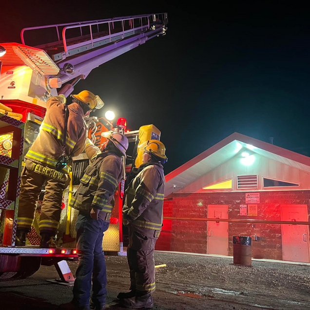 Firefighters in yellow gear stand beside a ladder truck outside a building at night.