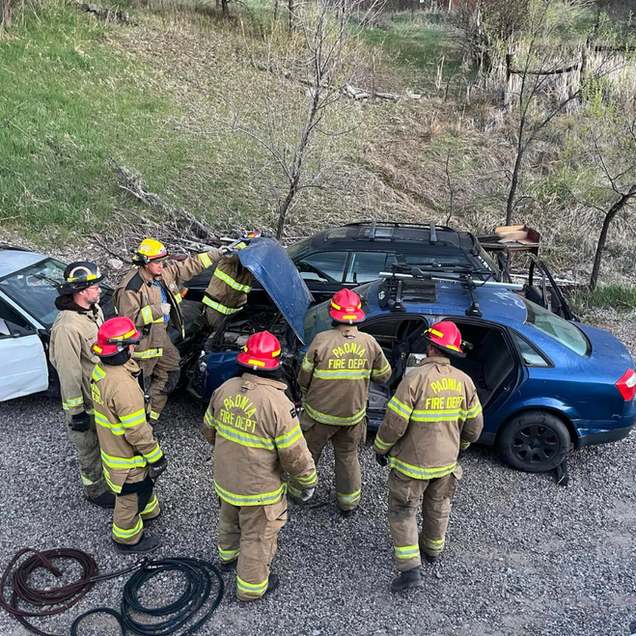 Firefighters in yellow gear gather around a blue car on a gravel roadside after a crash