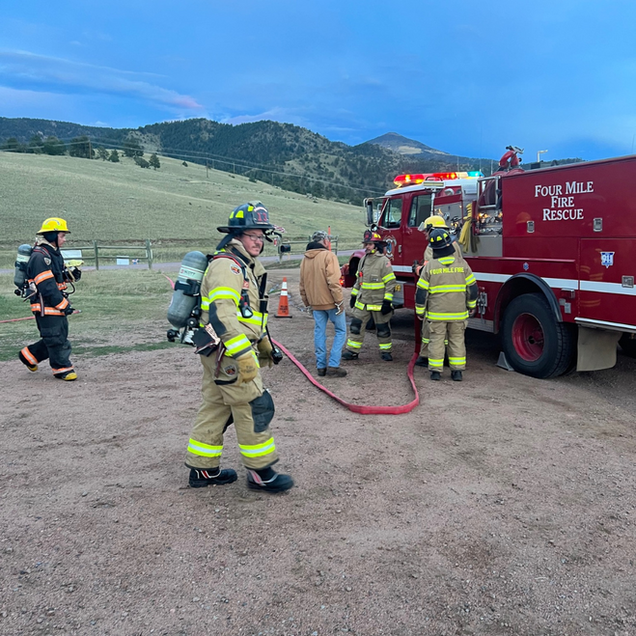 Firefighters in yellow gear stand beside a red rescue truck in a rural field with mountains behind them.