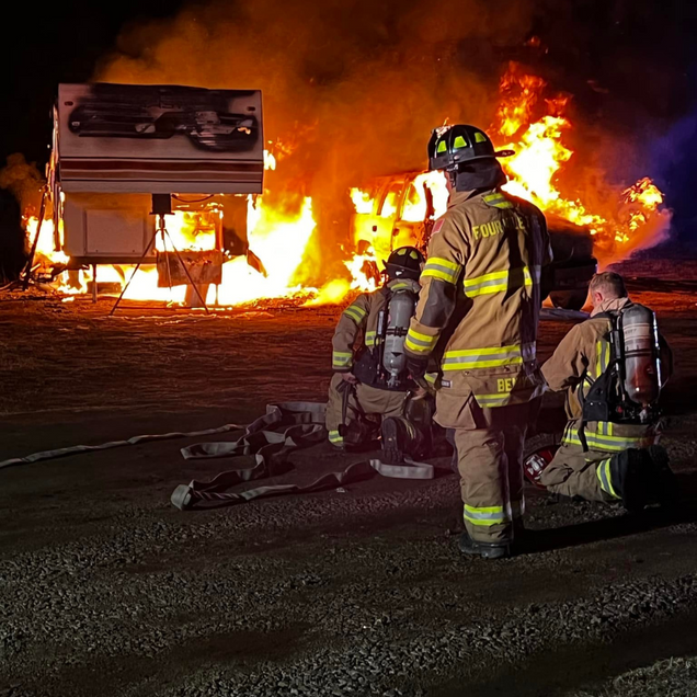 Firefighters kneel by a blaze near a truck at night, with flames and thick smoke filling the scene.