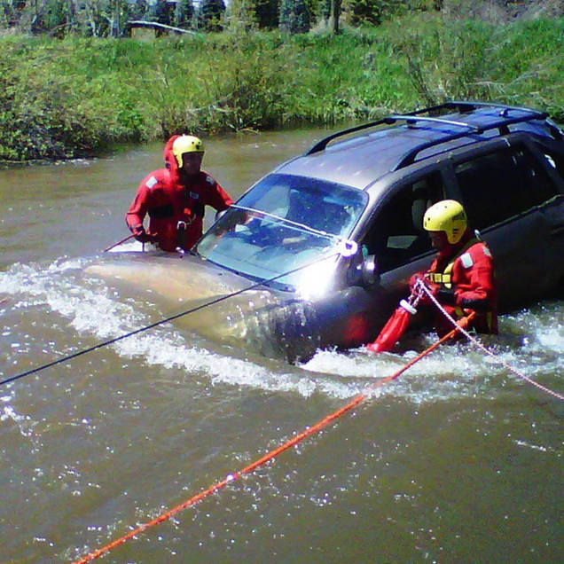 Two rescue workers in red gear use ropes to assist a stranded SUV in floodwater.