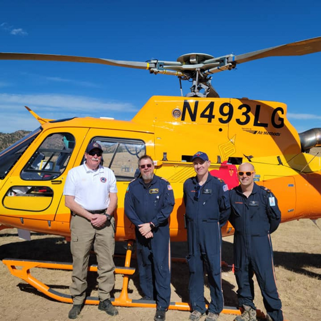 Four people stand beside a bright yellow helicopter on a sunny tarmac.