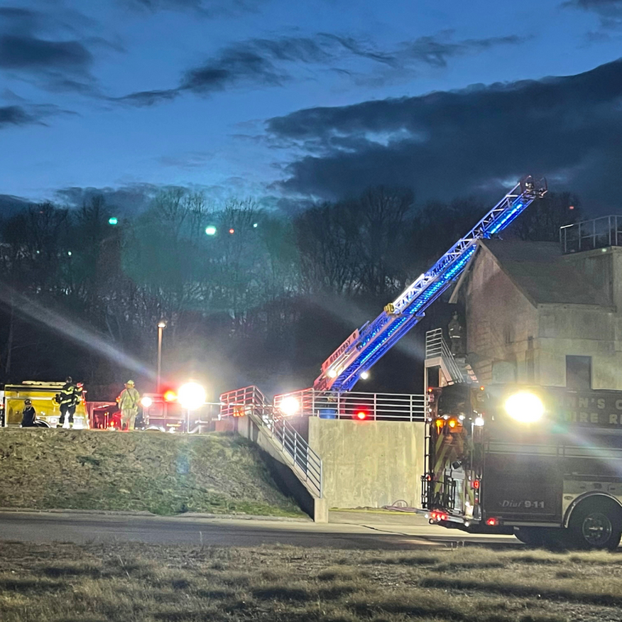 Fire trucks and firefighters at a nighttime roadside emergency, with a ladder extended toward a hillside building.