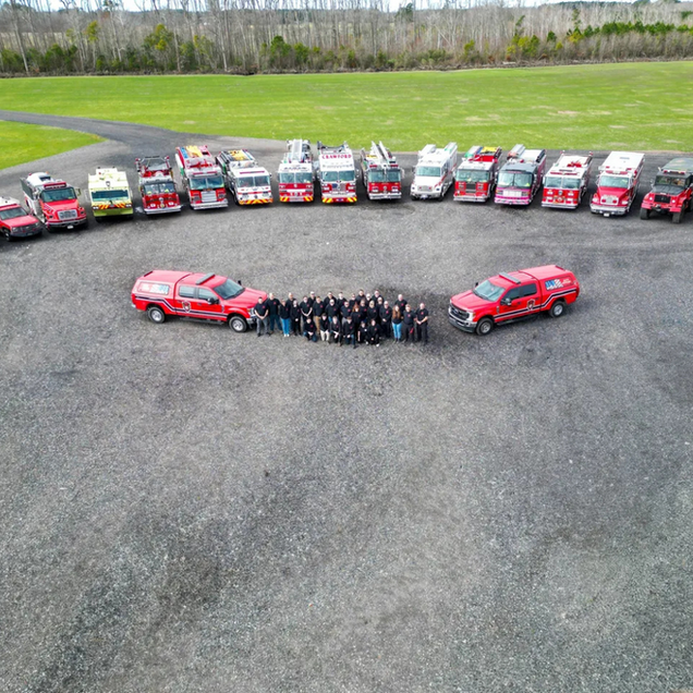 Aerial view of a group standing between two red cars, with a row of colorful cars behind them on a paved lot.