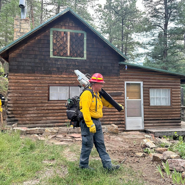 Firefighter in yellow gear standing outside a rustic cabin with a backpack leaf blower.