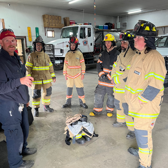 Firefighters in gear listening in a fire station garage beside a truck and equipment.