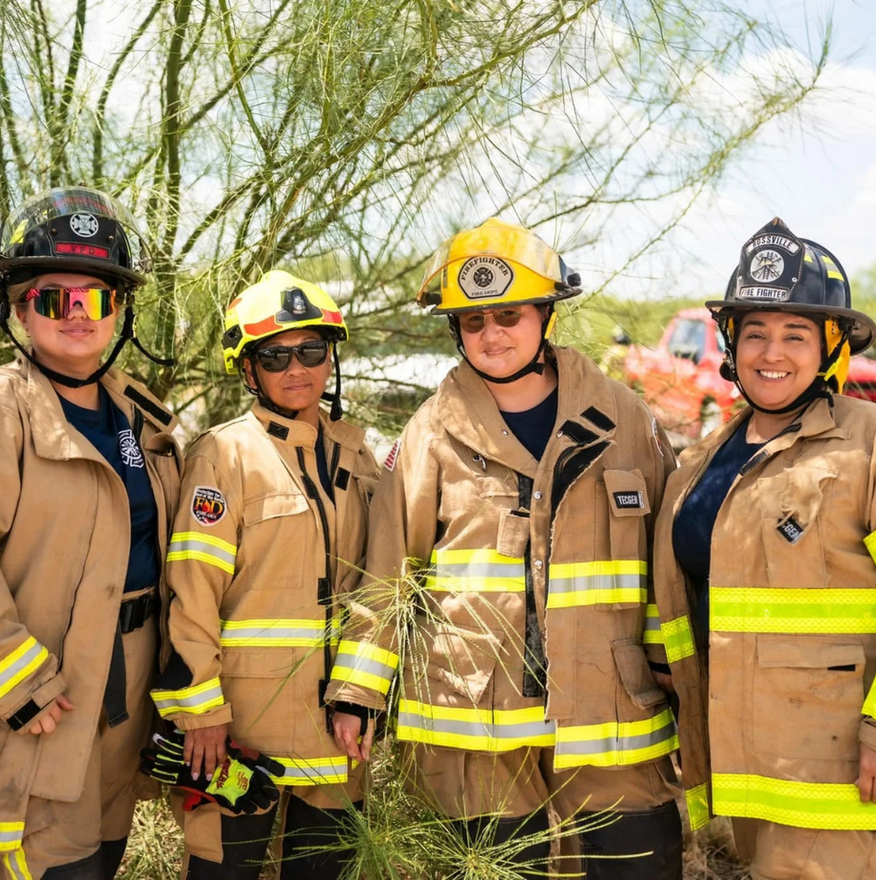 Four firefighters in tan gear and helmets standing outdoors, smiling at the camera.