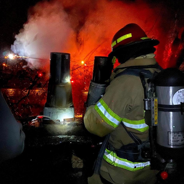 Firefighter with oxygen tank battling a blaze at night, bright orange flames and smoke in the background