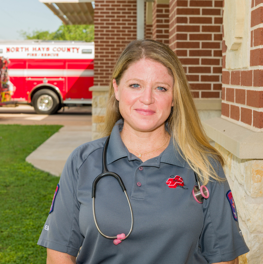 Paramedic in gray uniform stands outside a brick building with a red ambulance in the background.