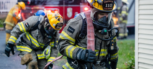 Firefighters in yellow gear crouch near a smoky house, with a fire truck flashing red lights behind them.