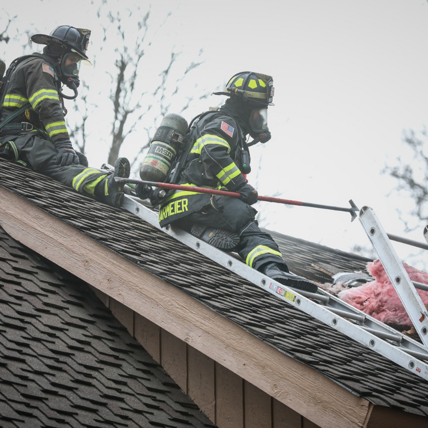 Two firefighters on a roof using a ladder and hose in a smoky scene