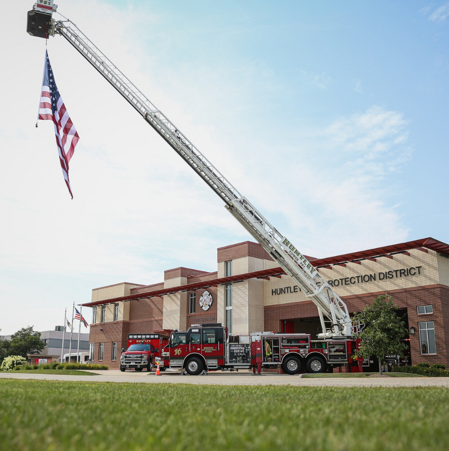 Fire truck ladder raised outside a brick fire station with an American flag flying nearby