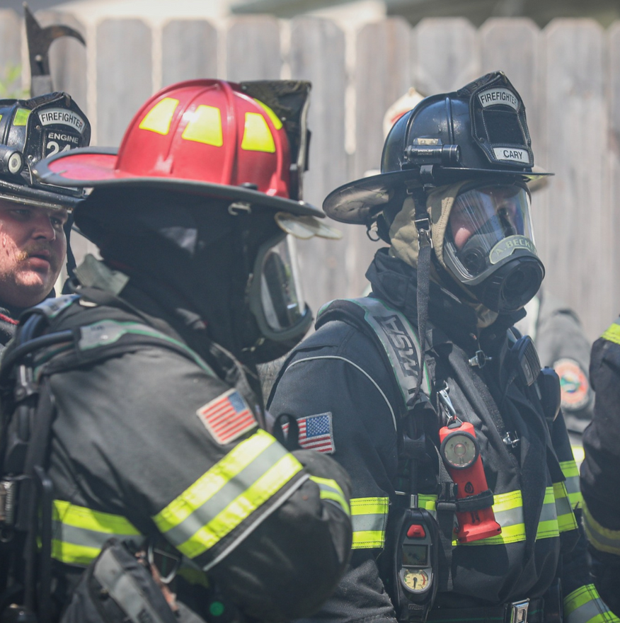 Firefighters in black gear and helmets stand together, one wearing a red helmet and face shield.