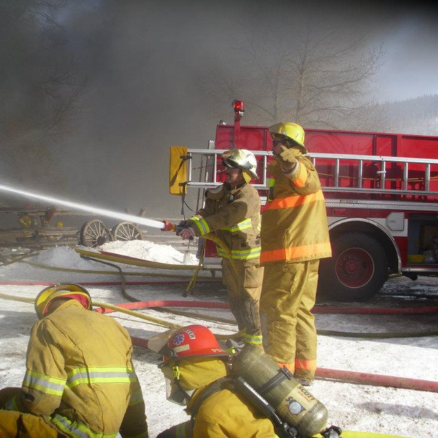 Firefighters in yellow gear spray water at a smoky scene beside a red fire truck.