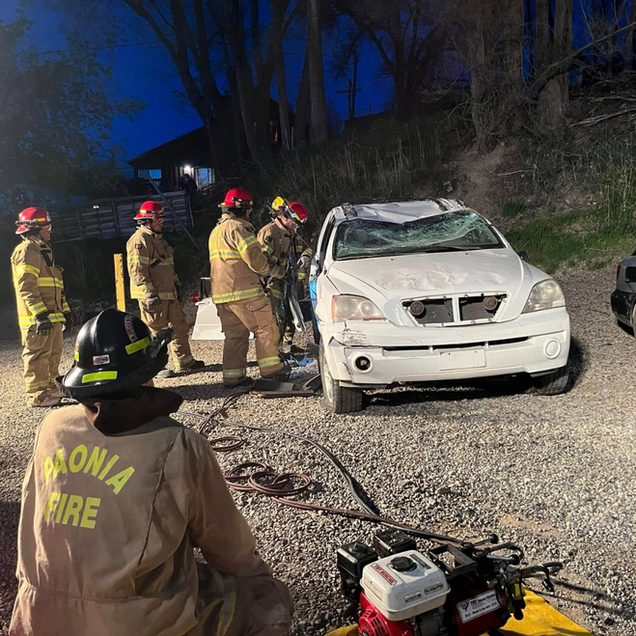 Firefighters at night inspect a wrecked white SUV on a gravel roadside.