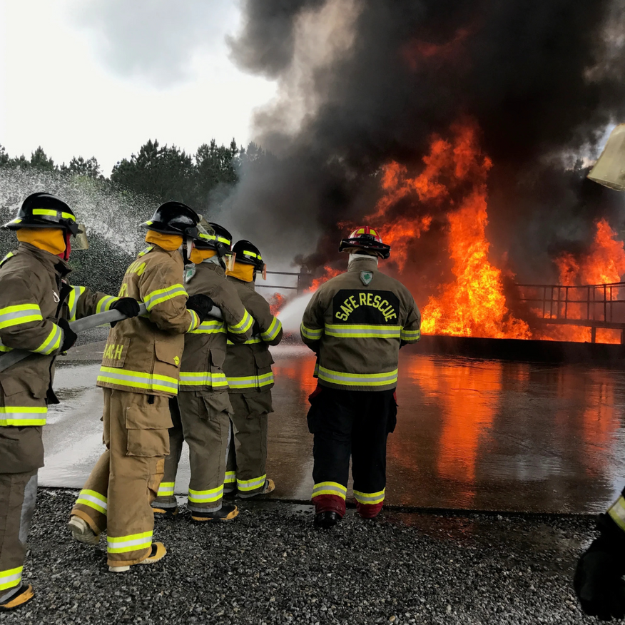 Firefighters in yellow gear face a large building fire with thick black smoke and bright flames.