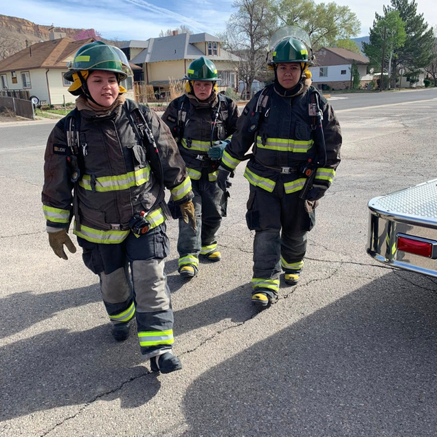 Three firefighters in gear walking down a residential street beside an emergency vehicle
