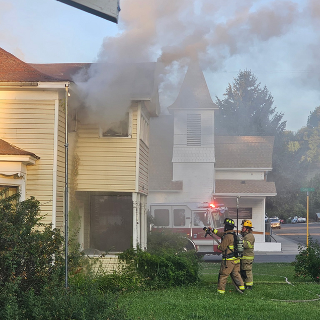 Firefighters battling smoke from a townhouse fire with flames near the roof and red emergency lights visible.