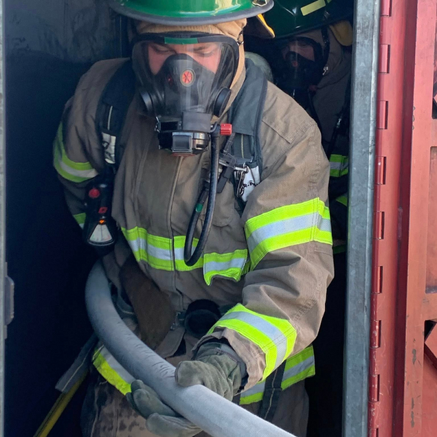 Firefighter in tan gear and mask holding a hose inside a doorway.
