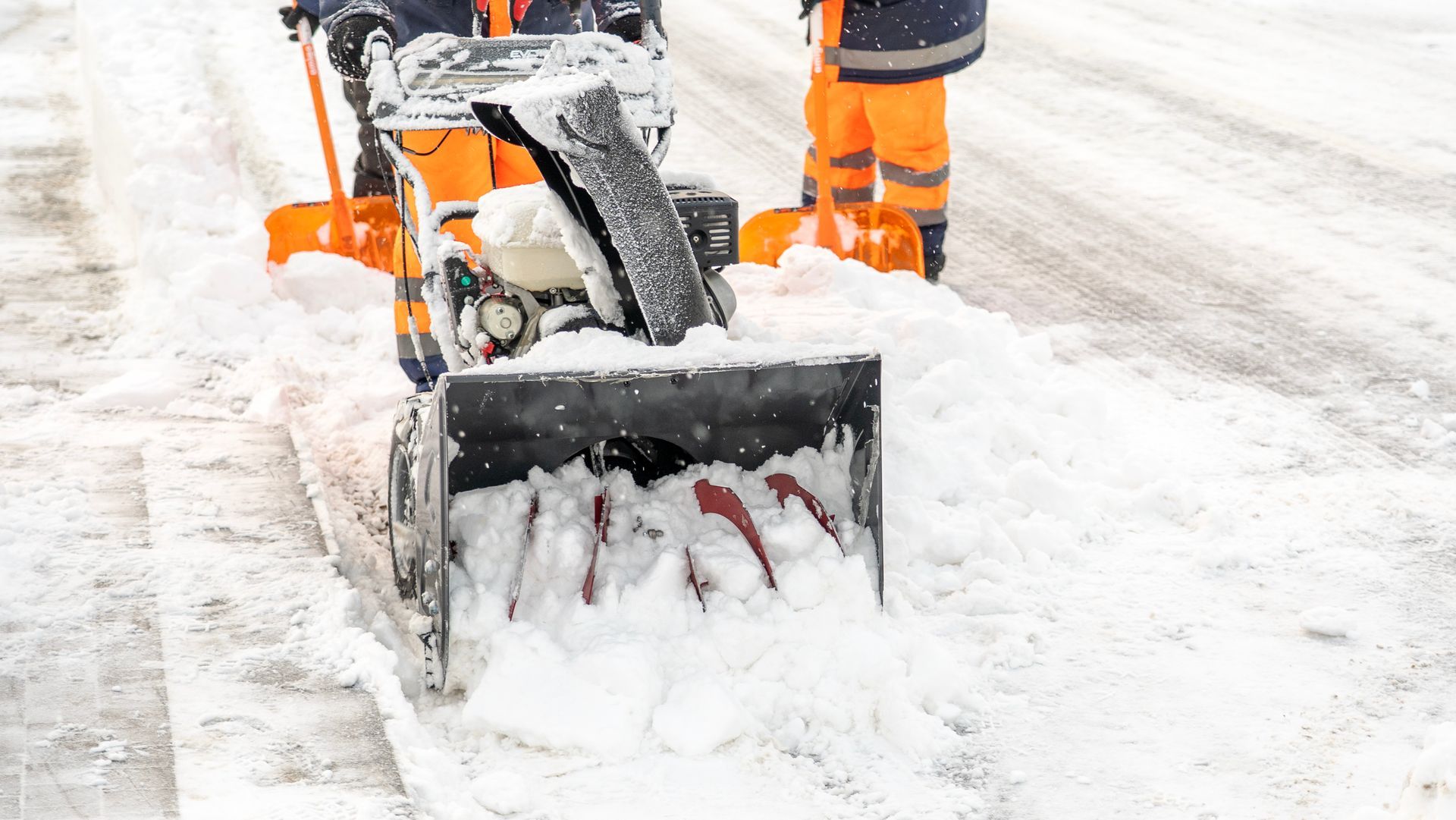 Snowblower clearing snow on a sidewalk, with worker’s orange boots visible behind it.