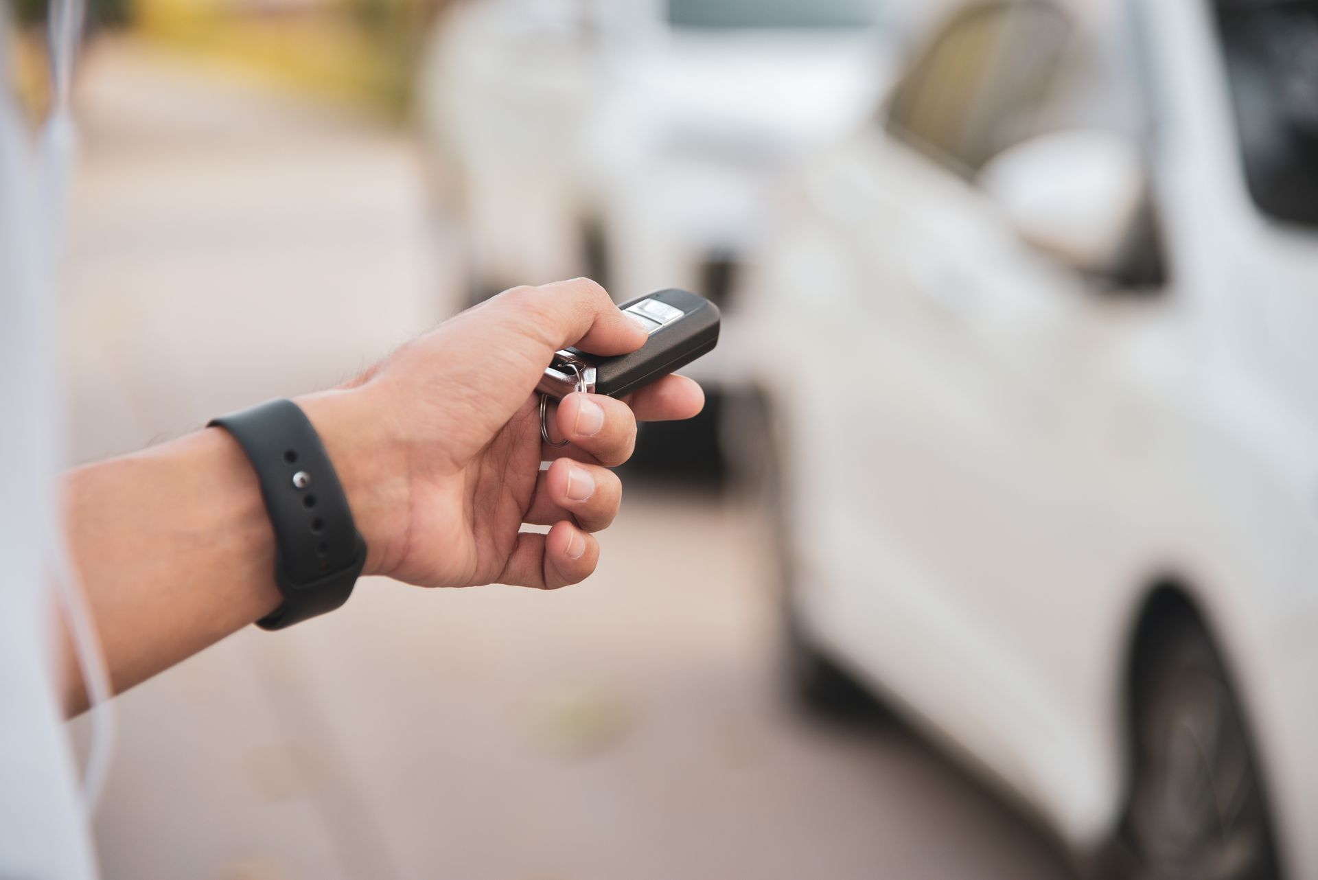 Hand holding a car key fob beside a white car on a street Hand holding a car key fob beside a white car on a street