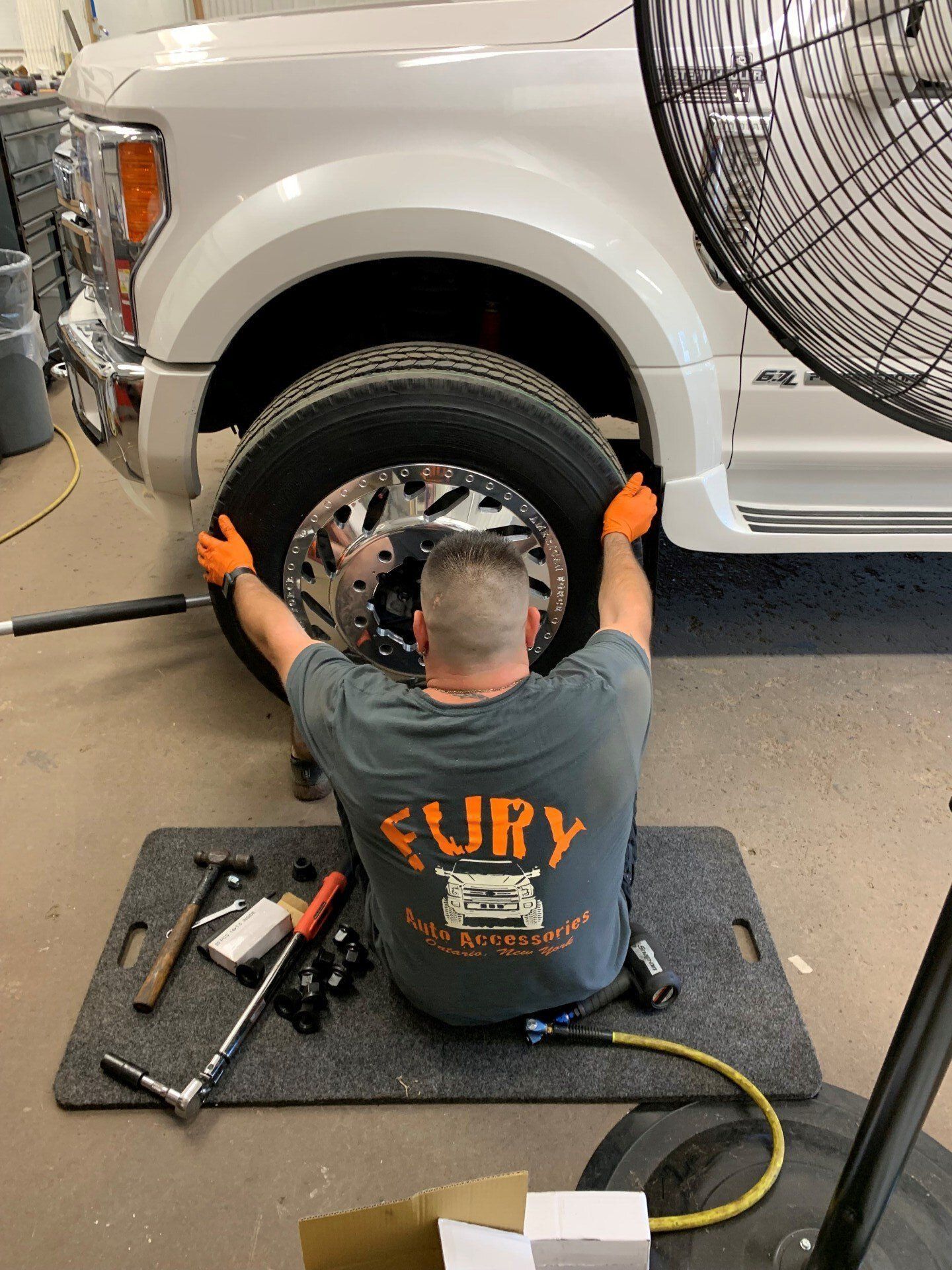 Mechanic seated under white SUV, working on front wheel with tools on a mat, orange gloves on