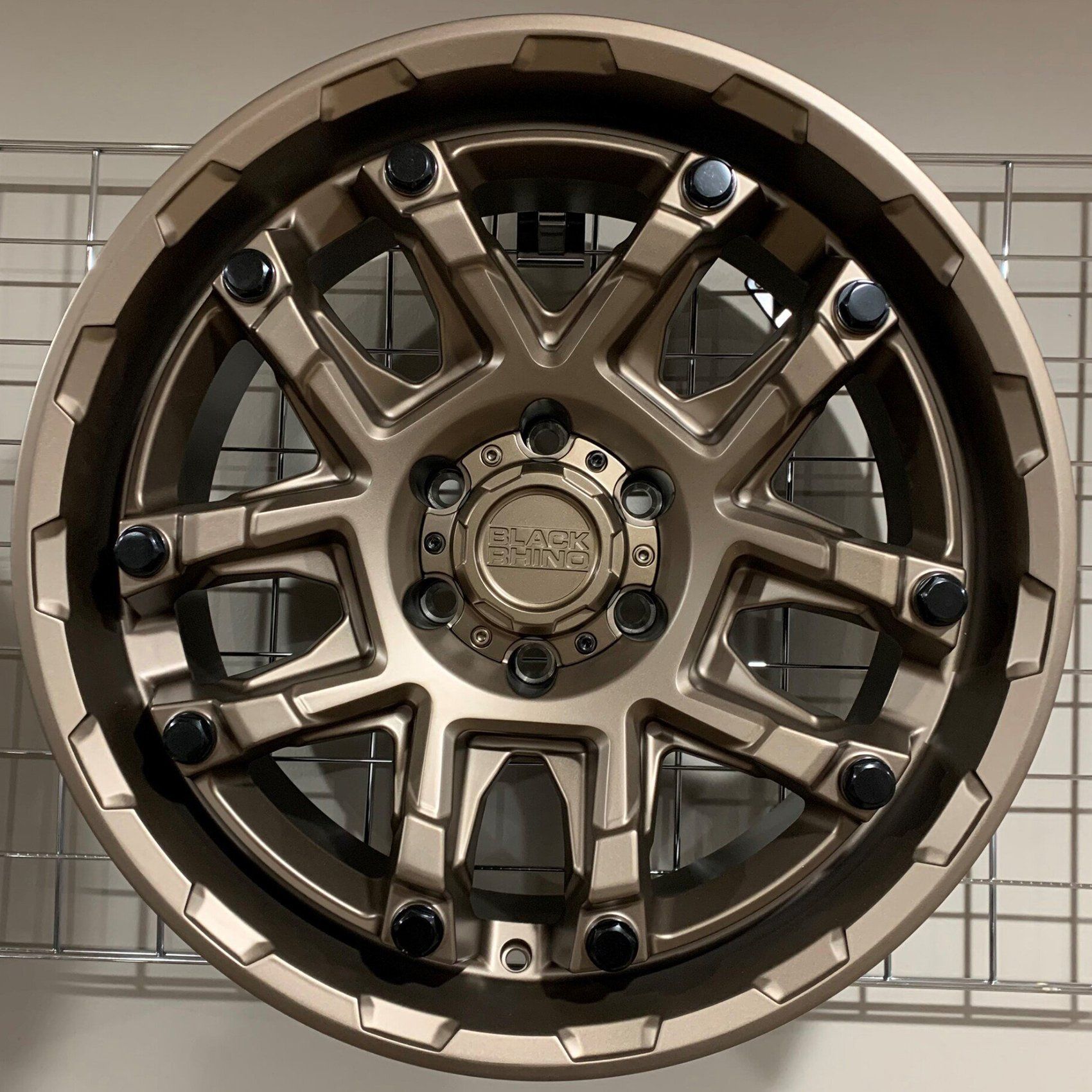 Bronze off-road wheel rim with black accents and blue bolts, shown front-on against a wall.