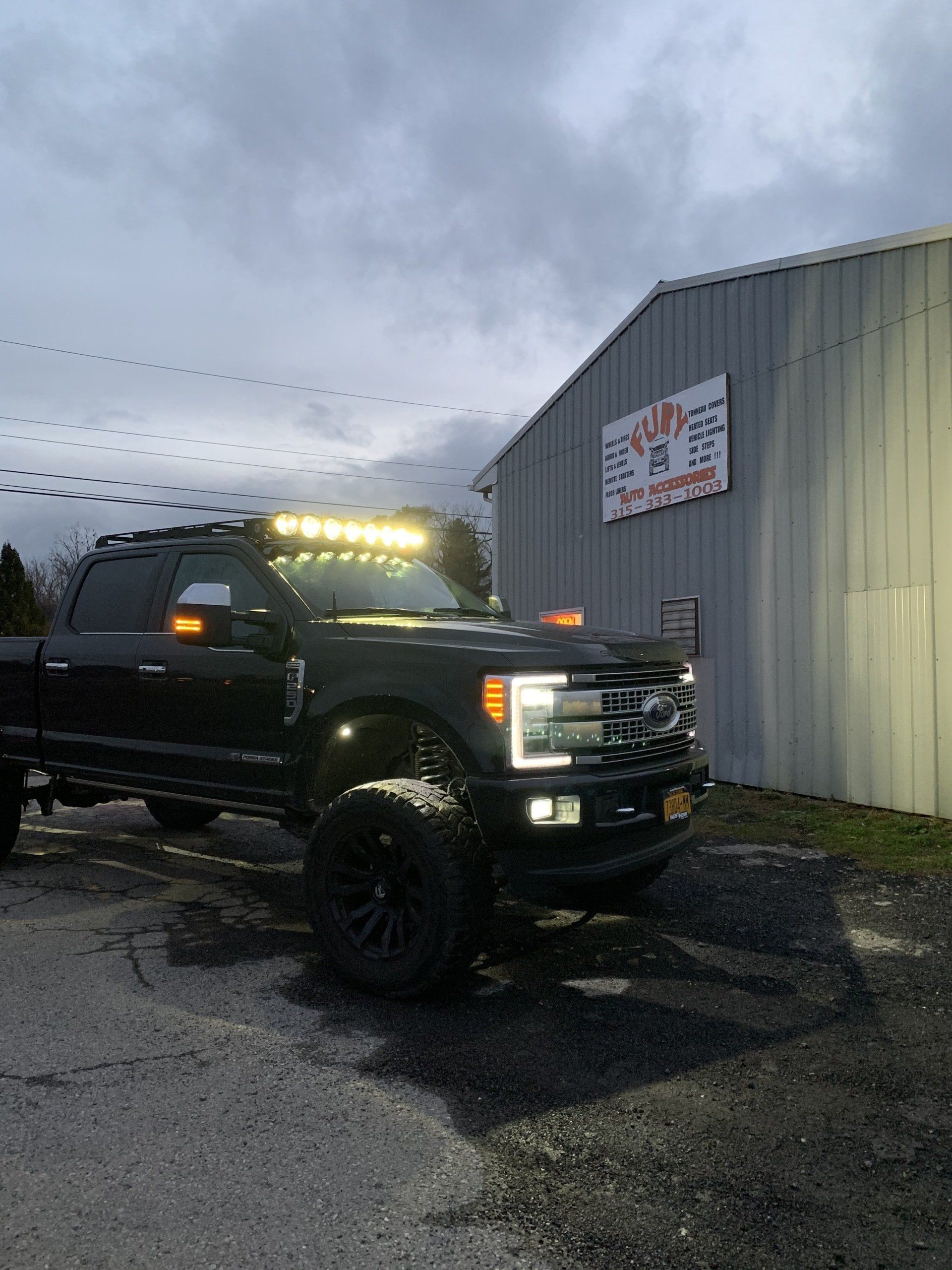 Black lifted pickup truck parked beside a metal building at dusk, lights on and wet pavement reflecting.