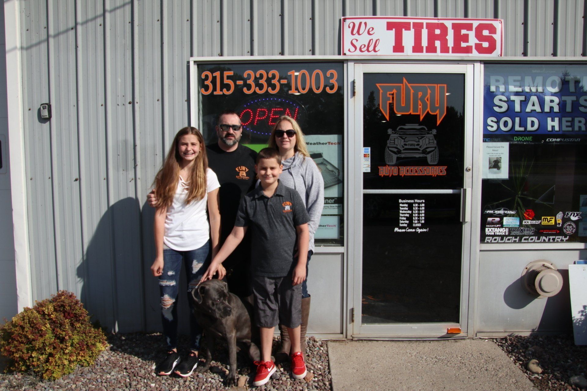 Four people standing outside a tire shop with a red-and-white sign and storefront window.