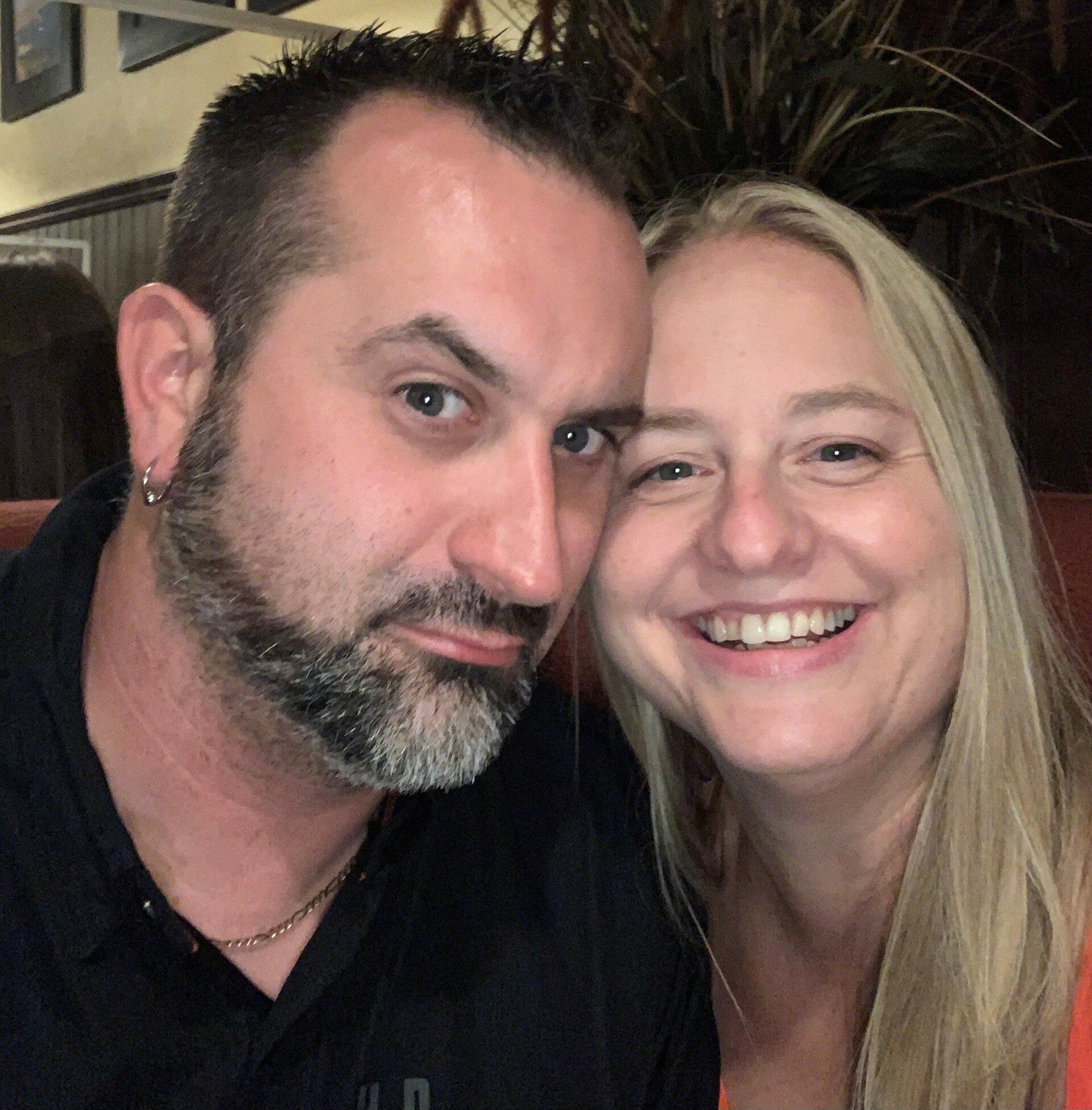 Two people smiling closely together indoors, one with a beard and the other with long blonde hair.