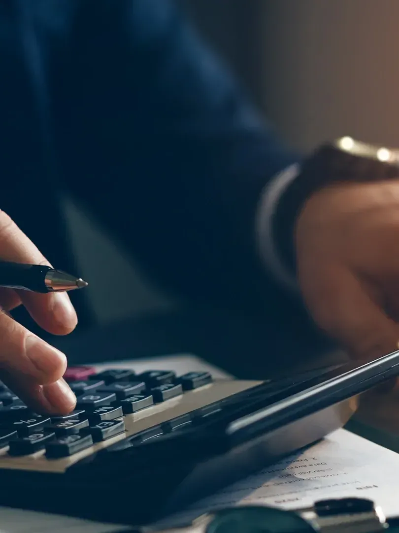 Hands calculating on a calculator beside a pen and paperwork in a dim office setting