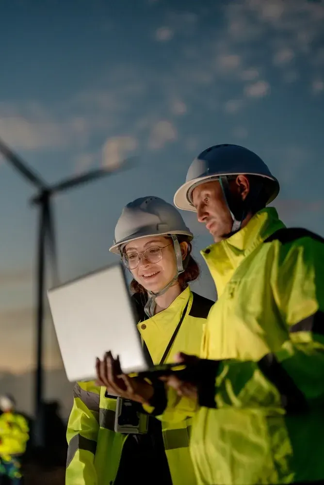Two engineers in neon safety jackets review a laptop near wind turbines at dusk.