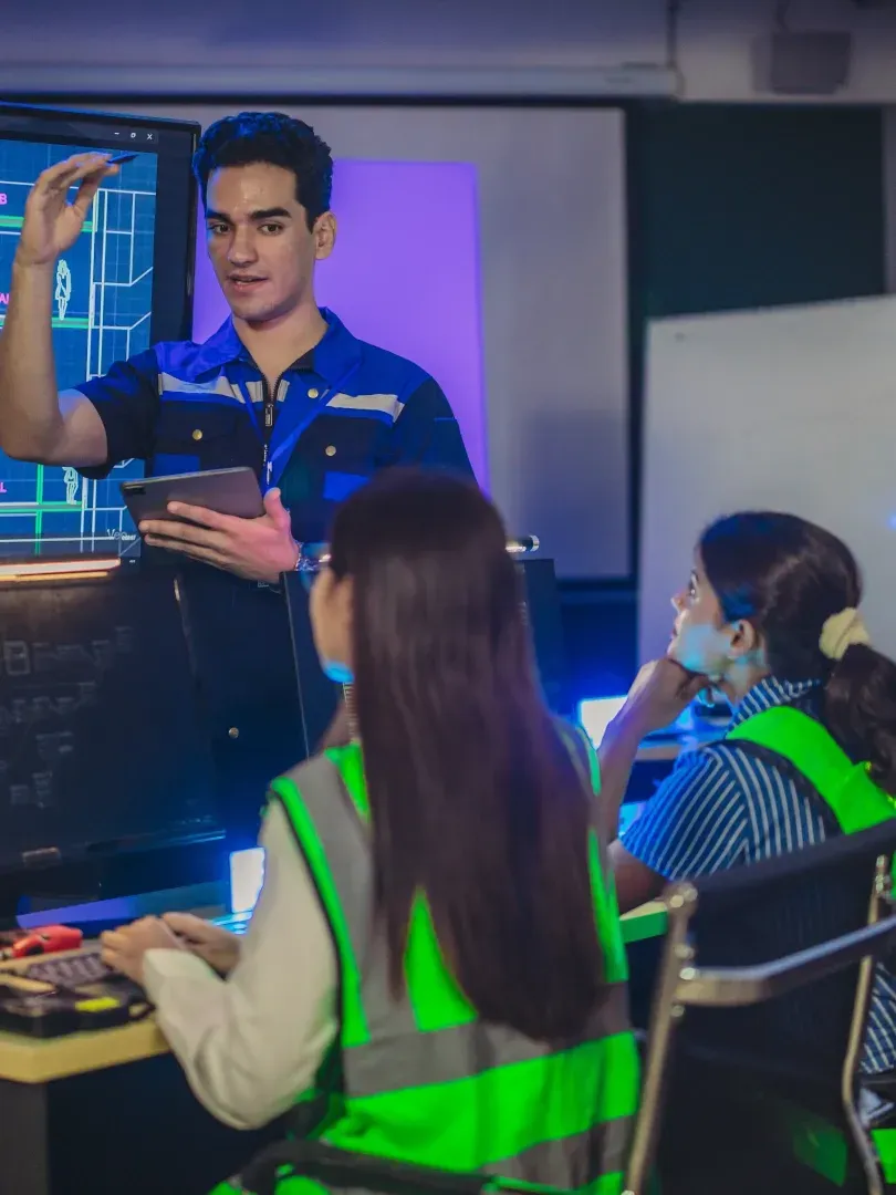 Instructor presenting to seated learners in a classroom, with one person using a tablet and safety vests visible.