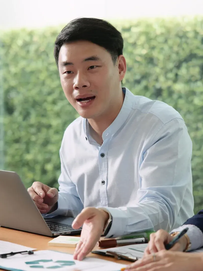 Person speaking in a meeting beside a laptop, with notebooks and pens on a desk.
